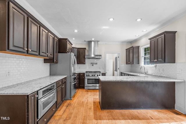 kitchen featuring a peninsula, a sink, ornamental molding, wall chimney range hood, and appliances with stainless steel finishes