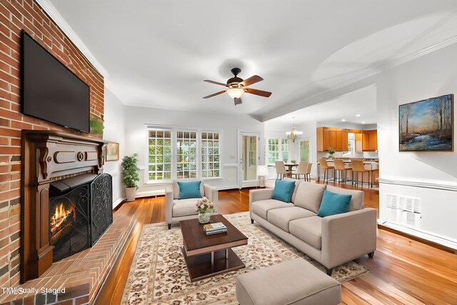 living area with ceiling fan with notable chandelier, a fireplace, crown molding, light wood-type flooring, and visible vents