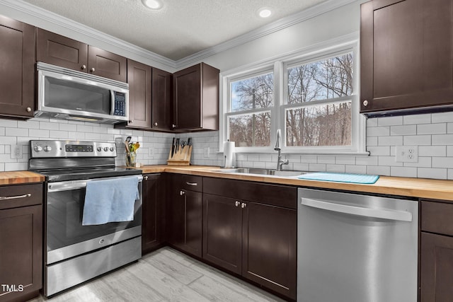 kitchen featuring crown molding, dark brown cabinetry, butcher block counters, stainless steel appliances, and a sink