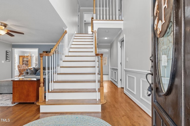 foyer with crown molding, stairs, ceiling fan with notable chandelier, wood finished floors, and a decorative wall