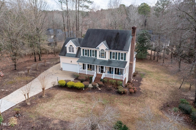 traditional-style home with a forest view, a shingled roof, concrete driveway, covered porch, and a chimney