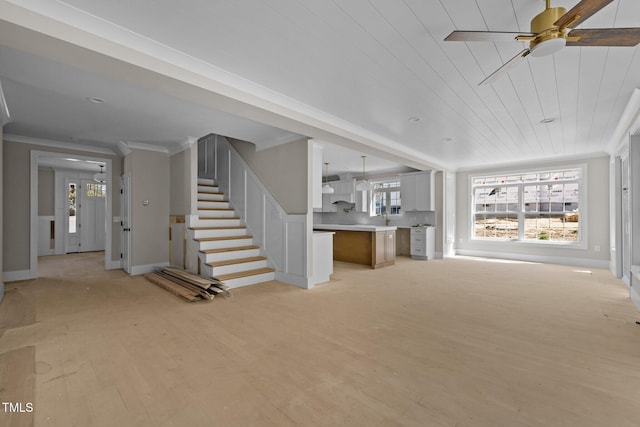 unfurnished living room featuring light wood-style flooring, crown molding, baseboards, ceiling fan, and stairs