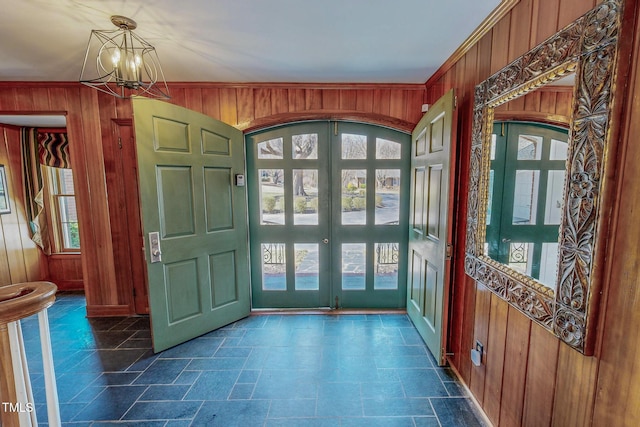 foyer featuring stone finish flooring, french doors, an inviting chandelier, wood walls, and crown molding