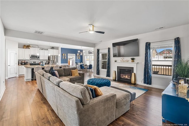 living room with ceiling fan with notable chandelier, visible vents, baseboards, hardwood / wood-style floors, and a glass covered fireplace