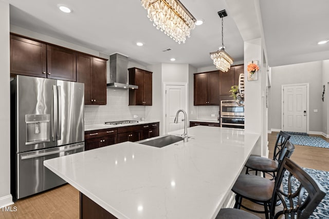 kitchen with light wood-style flooring, a sink, stainless steel appliances, an inviting chandelier, and wall chimney exhaust hood