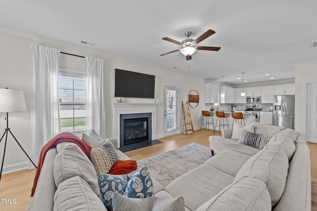 living room featuring light wood-style flooring, a fireplace with flush hearth, a ceiling fan, visible vents, and crown molding