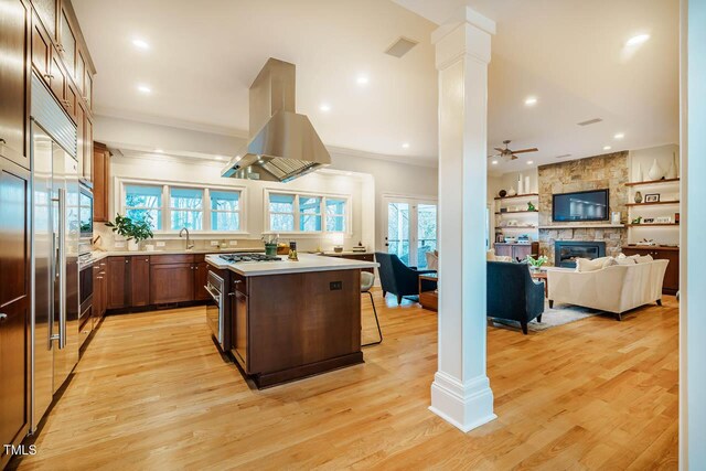 kitchen with a center island, light countertops, decorative columns, a stone fireplace, and island range hood