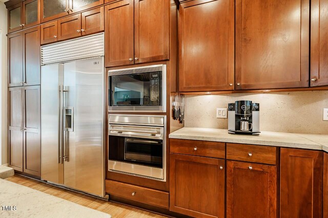 kitchen with backsplash, light wood-style floors, brown cabinetry, glass insert cabinets, and built in appliances