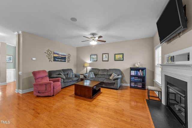 living area featuring baseboards, a fireplace with flush hearth, ceiling fan, and light wood finished floors
