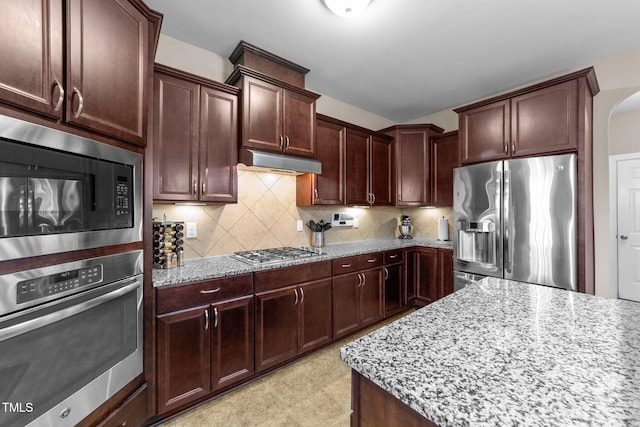 kitchen with under cabinet range hood, light stone counters, appliances with stainless steel finishes, decorative backsplash, and dark brown cabinets