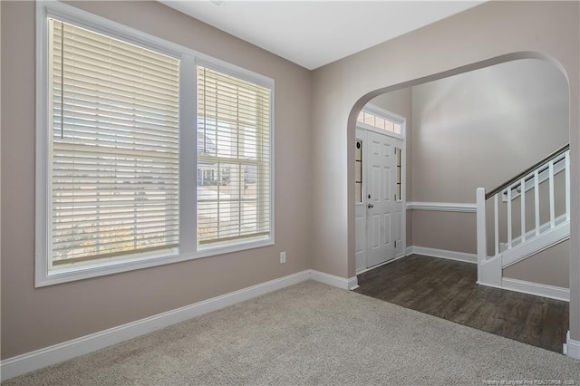 foyer featuring arched walkways, stairway, baseboards, and dark colored carpet