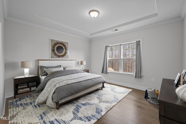 bedroom featuring wood finished floors, visible vents, baseboards, a tray ceiling, and crown molding