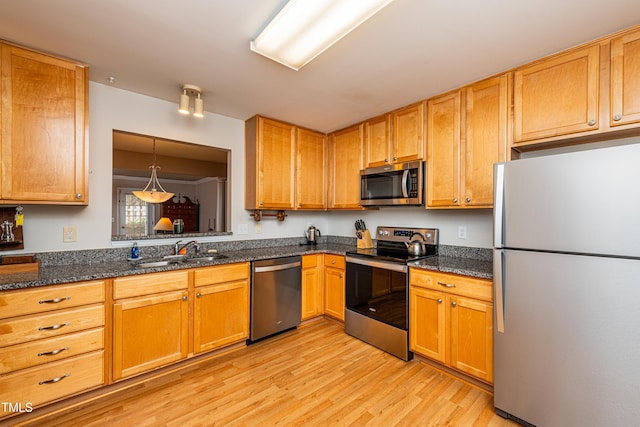 kitchen featuring light wood finished floors, stainless steel appliances, hanging light fixtures, a sink, and dark stone countertops