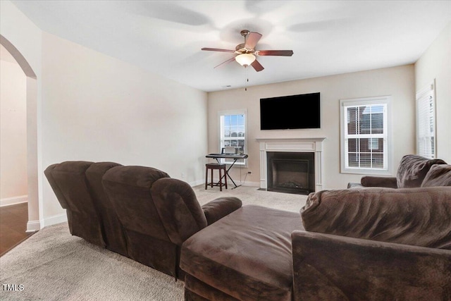 living area with arched walkways, light colored carpet, a fireplace with flush hearth, ceiling fan, and baseboards