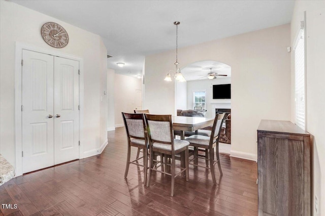 dining room featuring arched walkways, ceiling fan with notable chandelier, dark wood-type flooring, a fireplace, and baseboards