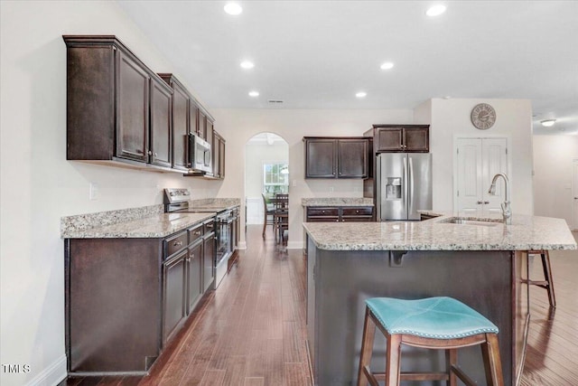 kitchen with arched walkways, wood finished floors, stainless steel appliances, dark brown cabinets, and a sink