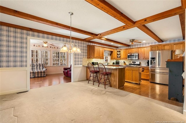 kitchen featuring brown cabinets, stainless steel appliances, light colored carpet, beamed ceiling, and wallpapered walls