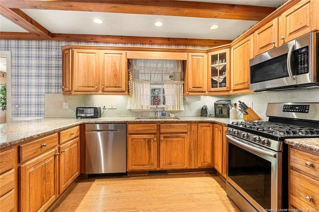 kitchen featuring wallpapered walls, light stone counters, a sink, stainless steel appliances, and beam ceiling