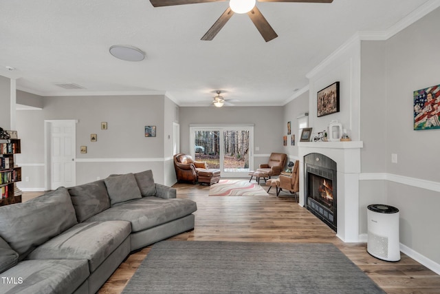 living area with baseboards, a ceiling fan, a tiled fireplace, ornamental molding, and wood finished floors