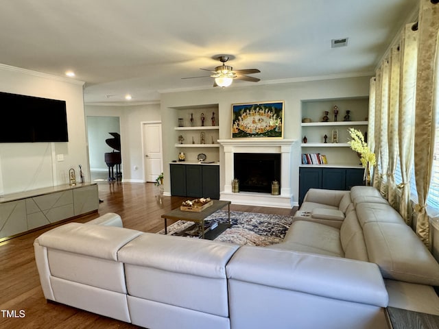 living room featuring a fireplace with raised hearth, built in shelves, wood finished floors, visible vents, and crown molding