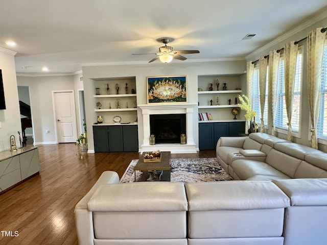 living area with a fireplace with raised hearth, built in shelves, dark wood-style flooring, baseboards, and crown molding