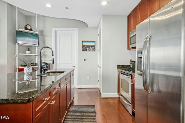 kitchen with baseboards, appliances with stainless steel finishes, dark stone countertops, wood finished floors, and a sink