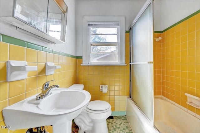 bathroom with tile walls, toilet, wainscoting, combined bath / shower with glass door, and a sink