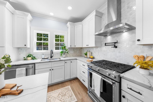 kitchen featuring appliances with stainless steel finishes, a sink, wall chimney exhaust hood, white cabinetry, and tasteful backsplash