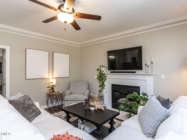 living area with a tiled fireplace, a ceiling fan, baseboards, and ornamental molding