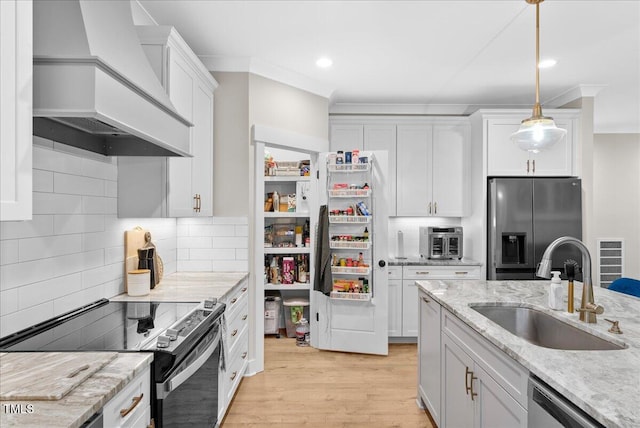 kitchen with light wood finished floors, appliances with stainless steel finishes, crown molding, premium range hood, and a sink