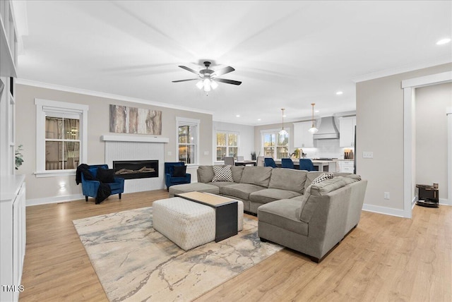 living area featuring baseboards, a ceiling fan, a glass covered fireplace, ornamental molding, and light wood-type flooring