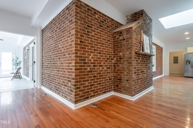 corridor featuring brick wall, baseboards, electric panel, light wood-style flooring, and a skylight