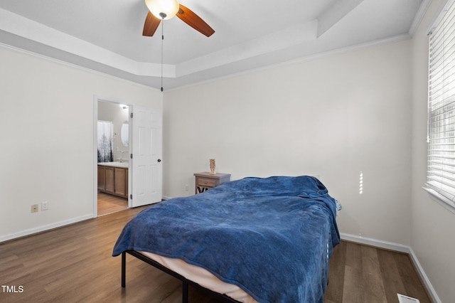 bedroom with wood finished floors, baseboards, ensuite bath, a tray ceiling, and crown molding