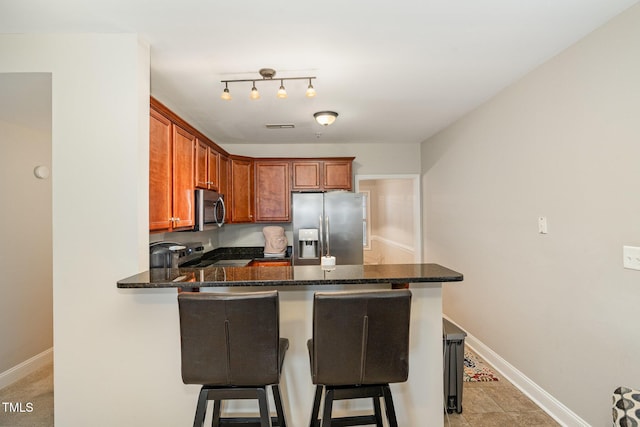 kitchen with a kitchen bar, stainless steel appliances, dark stone counters, a peninsula, and baseboards