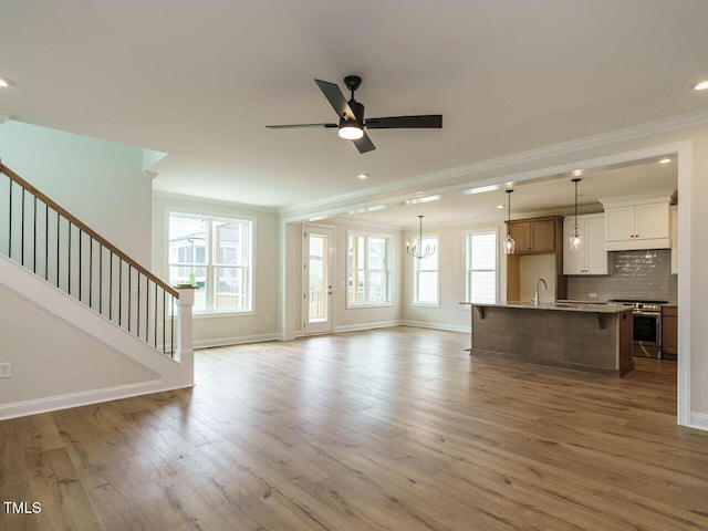 unfurnished living room with a wealth of natural light, stairway, light wood finished floors, and ceiling fan with notable chandelier