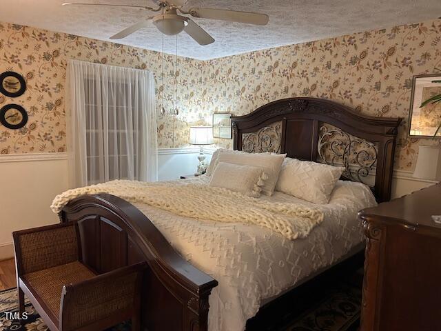 bedroom with a textured ceiling, ceiling fan, wainscoting, and wallpapered walls