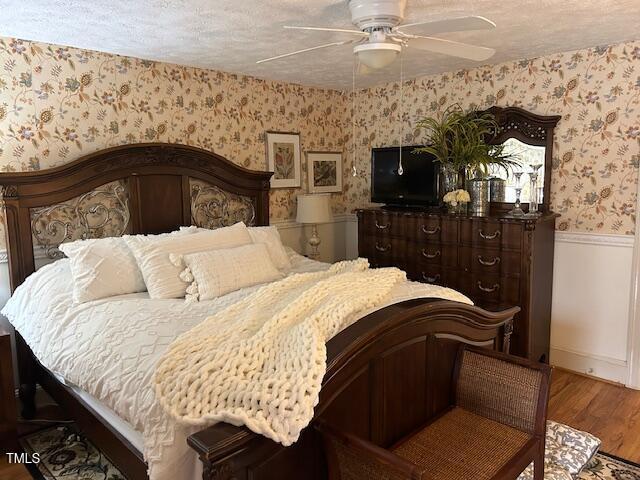 bedroom with wood finished floors, a wainscoted wall, wallpapered walls, ceiling fan, and a textured ceiling