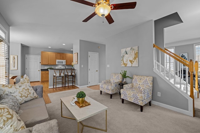 living room with stairway, light colored carpet, baseboards, and a ceiling fan