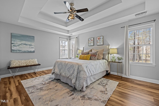 bedroom featuring visible vents, a tray ceiling, wood finished floors, baseboards, and ceiling fan