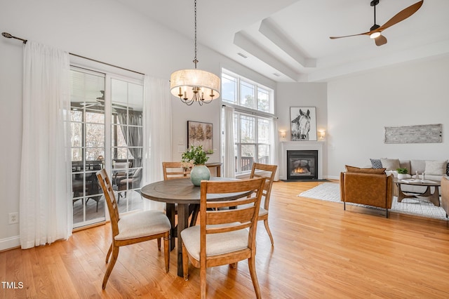 dining room featuring light wood-type flooring, ceiling fan with notable chandelier, a tray ceiling, a glass covered fireplace, and a high ceiling