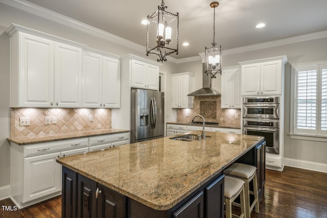 kitchen with a sink, wall chimney range hood, ornamental molding, appliances with stainless steel finishes, and a kitchen island with sink