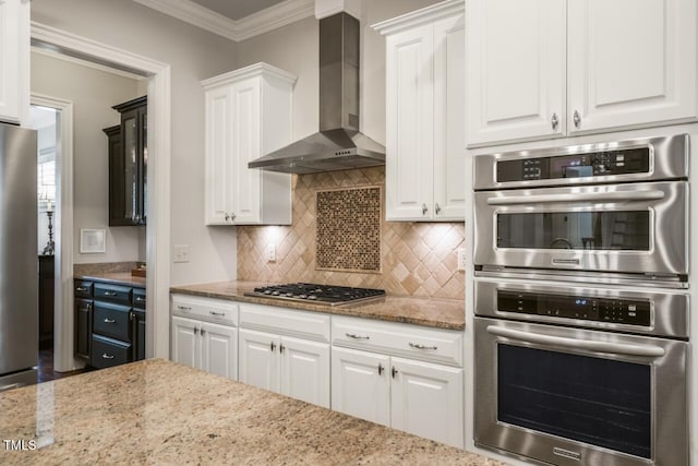 kitchen featuring light stone countertops, stainless steel appliances, decorative backsplash, white cabinetry, and wall chimney range hood