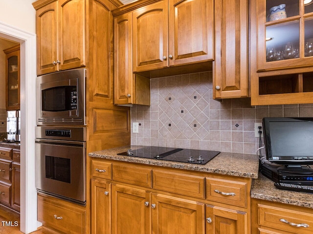 kitchen with light stone countertops, decorative backsplash, glass insert cabinets, appliances with stainless steel finishes, and brown cabinets