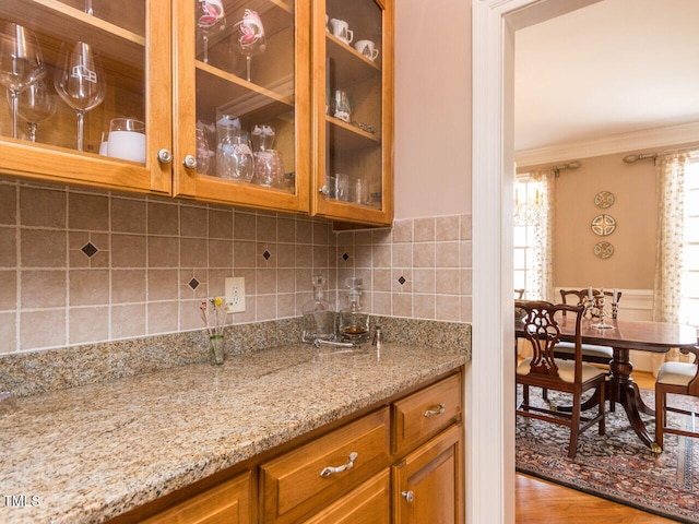 kitchen with wood finished floors, brown cabinetry, light stone countertops, glass insert cabinets, and crown molding