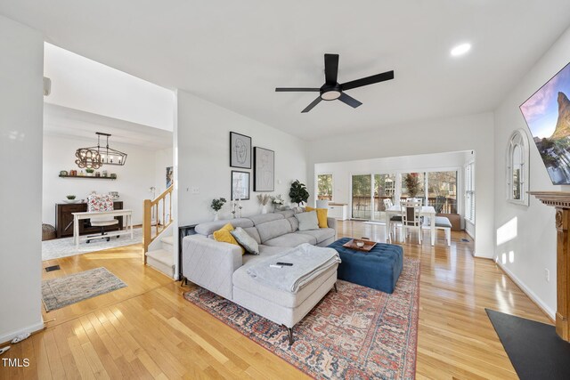 living room with light wood-type flooring, ceiling fan with notable chandelier, baseboards, and stairway