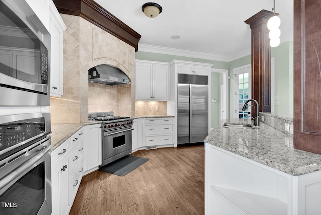 kitchen with high end appliances, wood finished floors, a sink, crown molding, and wall chimney exhaust hood