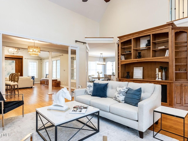 living area featuring baseboards, light wood-type flooring, ceiling fan with notable chandelier, high vaulted ceiling, and ornate columns