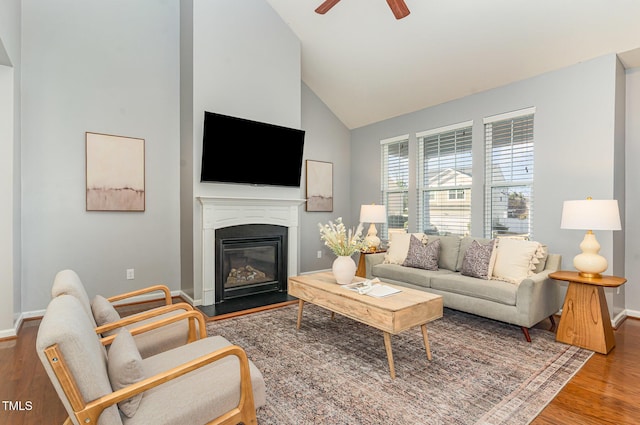 living area featuring high vaulted ceiling, a glass covered fireplace, wood finished floors, baseboards, and ceiling fan