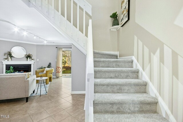 stairs with tile patterned floors, a warm lit fireplace, rail lighting, and crown molding