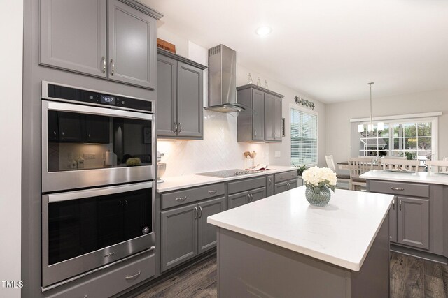 kitchen with black electric stovetop, gray cabinetry, double oven, wall chimney range hood, and a chandelier